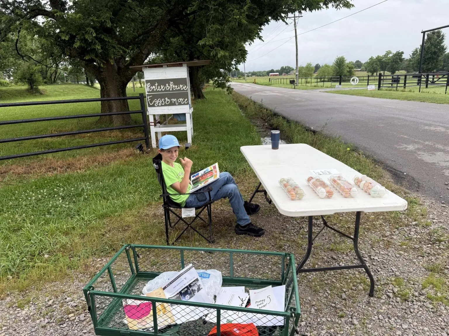 A child sits at the Porter & Prairie roadside egg stand beside a folding table of fresh eggs and a cooler.